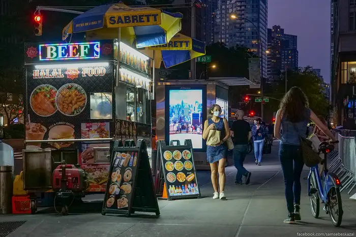A woman wearing a surgical mask walks past a street food vendor on a darkened NYC sidewalk.